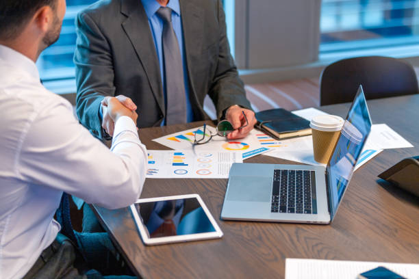 Businessmen shaking hands. Both are dressed in business clothing and there is paperwork with finance graphs and charts. There is also technology including a laptop computer and digital tablet